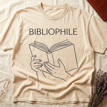 Ivory tee with two hands cradling an open book in cream and brown linework, "BIBLIOPHILE" in serif capitals above.