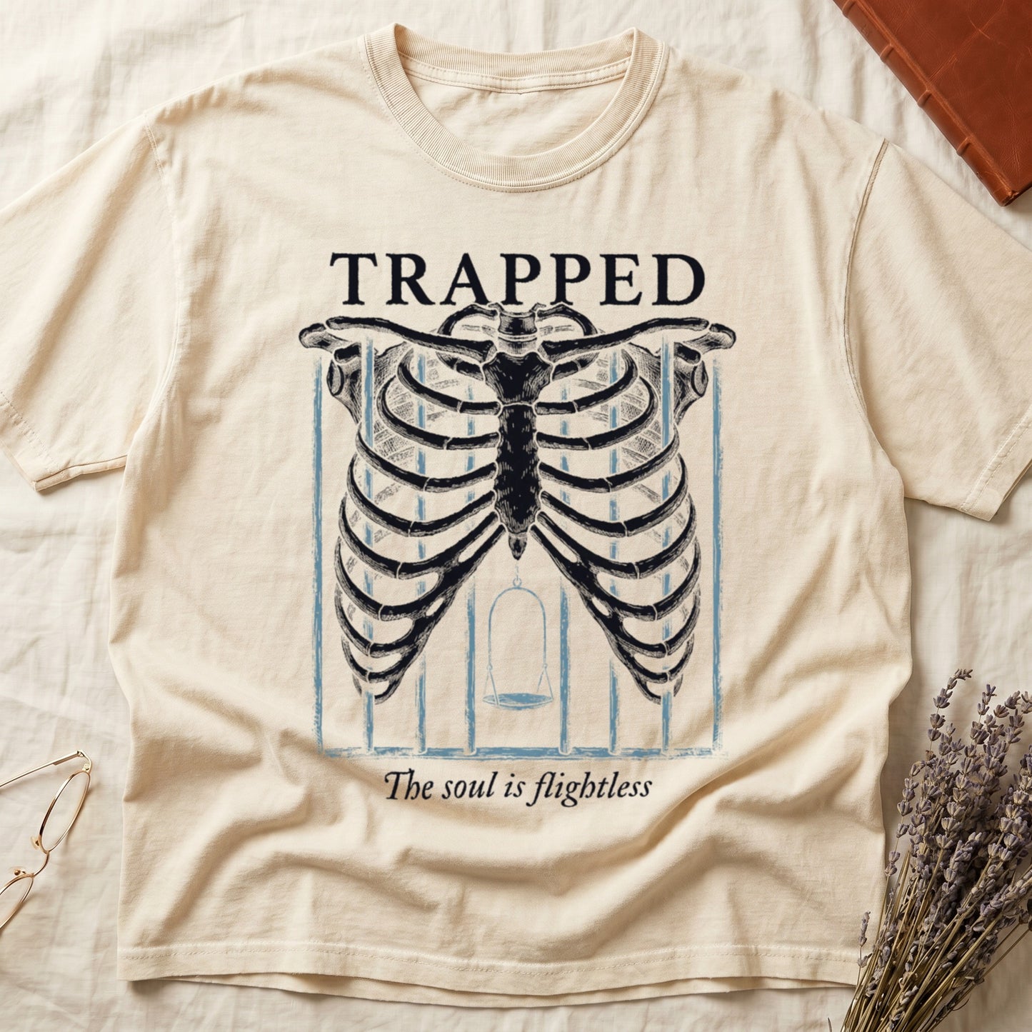 Ivory tee with anatomical ribcage on prison bars, bird swing inside, "TRAPPED" top, "The soul is flightless" italic below.
