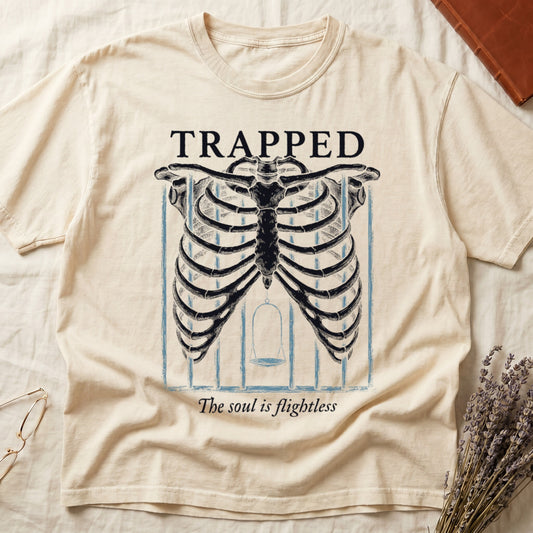 Ivory tee with anatomical ribcage on prison bars, bird swing inside, "TRAPPED" top, "The soul is flightless" italic below.