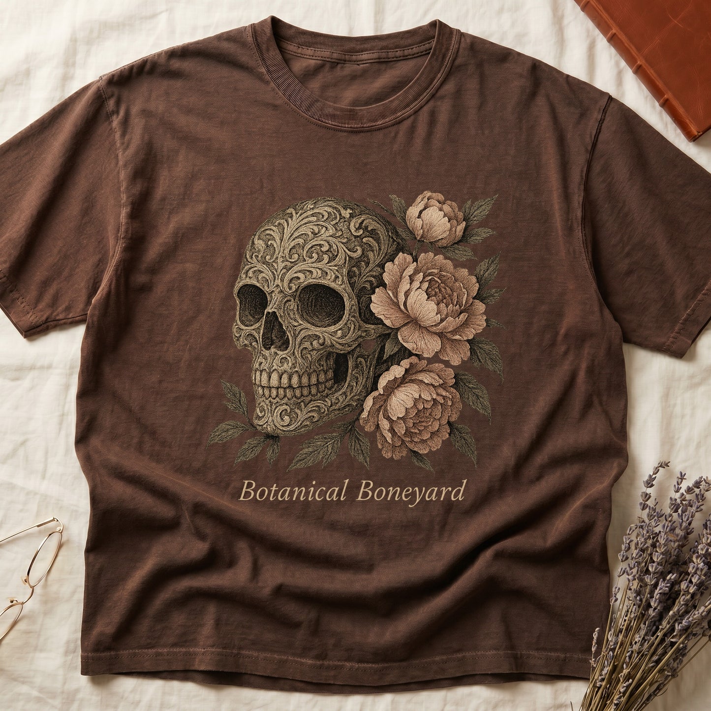 Espresso tee with filigree-engraved skull on left, peach-cream peonies on right, crossed bones below, "Botanical Boneyard."
