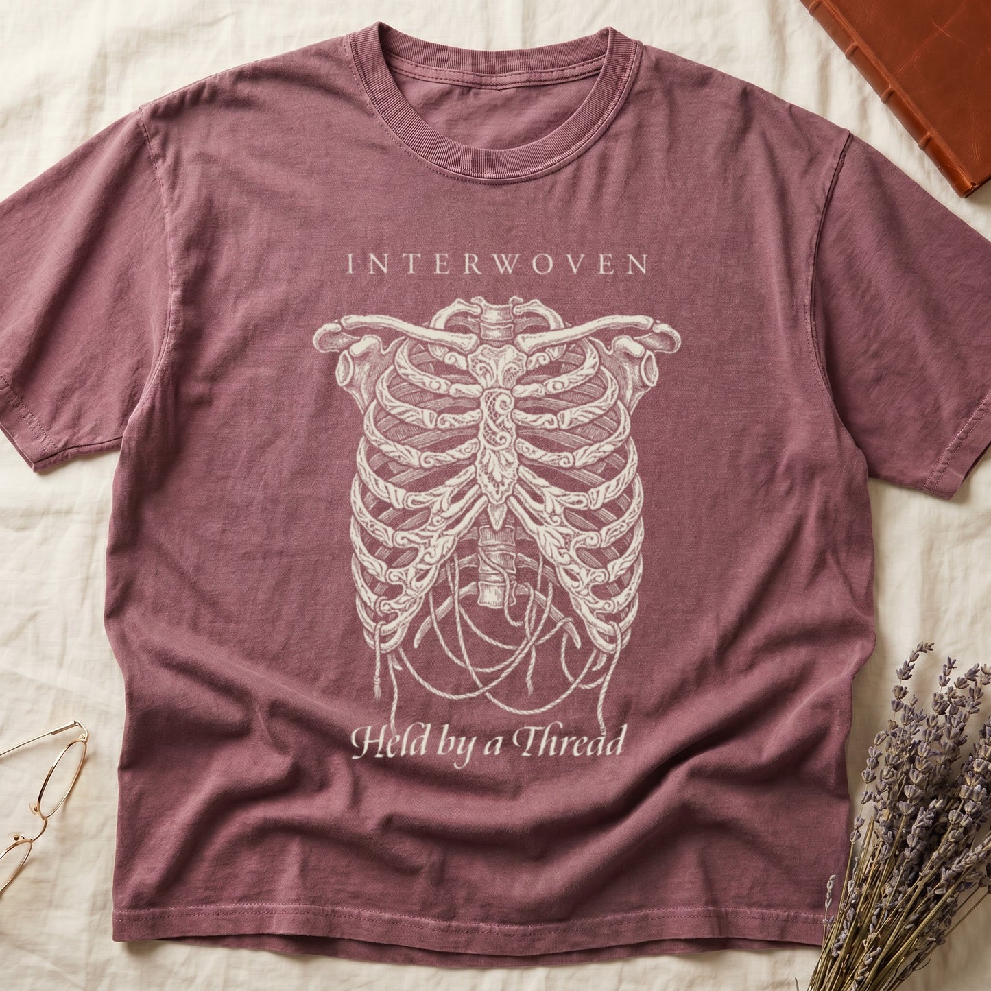 Dark Rose tee with cream skeletal ribcage suspended by trailing threads, "Held by a Thread" in serif on plate below.