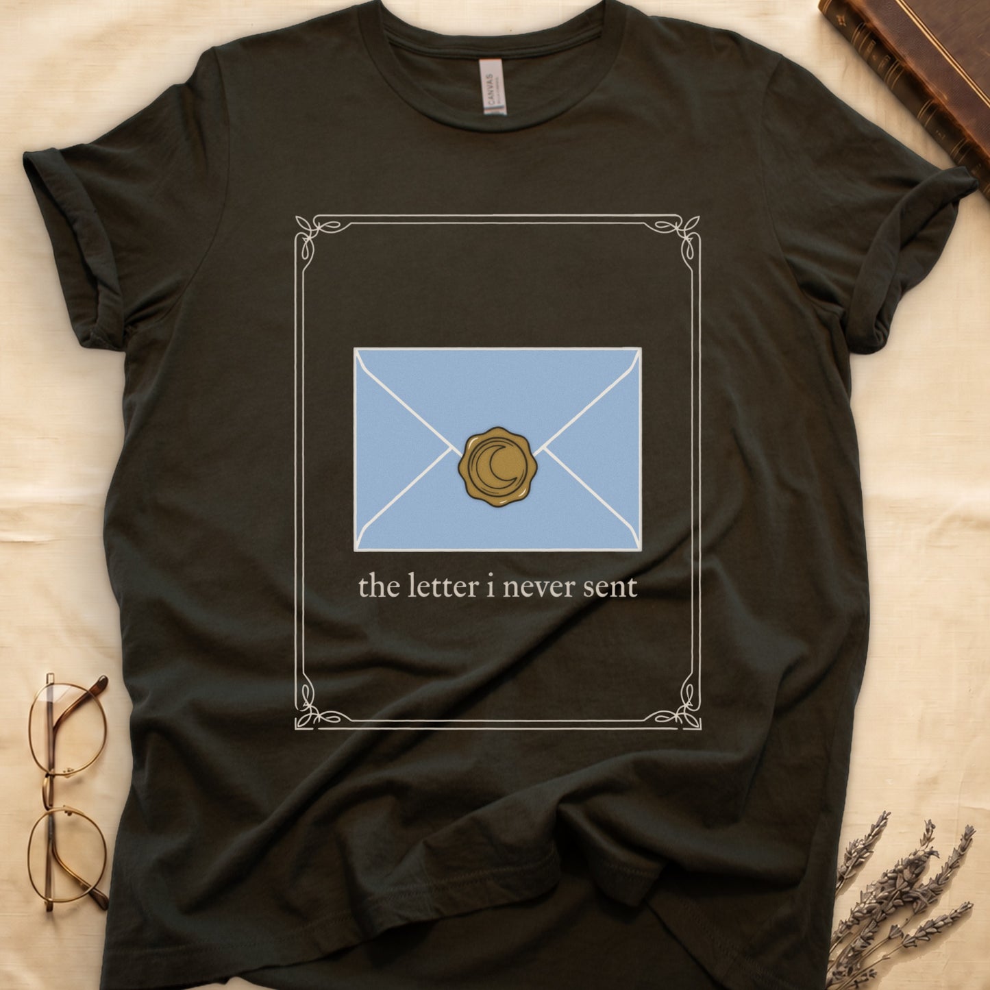 Military Green tee with pale blue envelope sealed with gold crescent moon wax seal, "the letter i never sent" cream serif below.