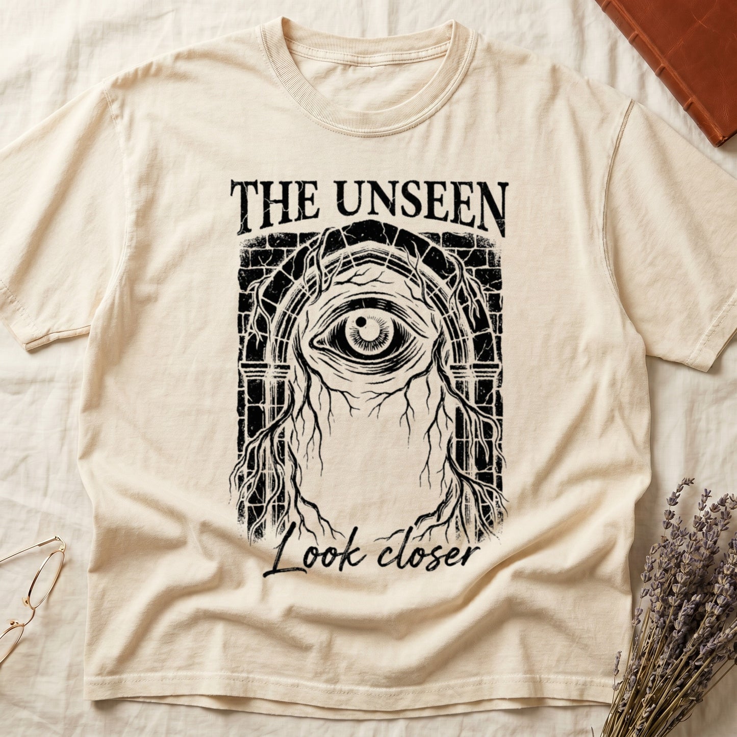 Ivory tee with Gothic stone arch, open eye, roots trailing, "THE UNSEEN" top, "Look closer" cursive below.
