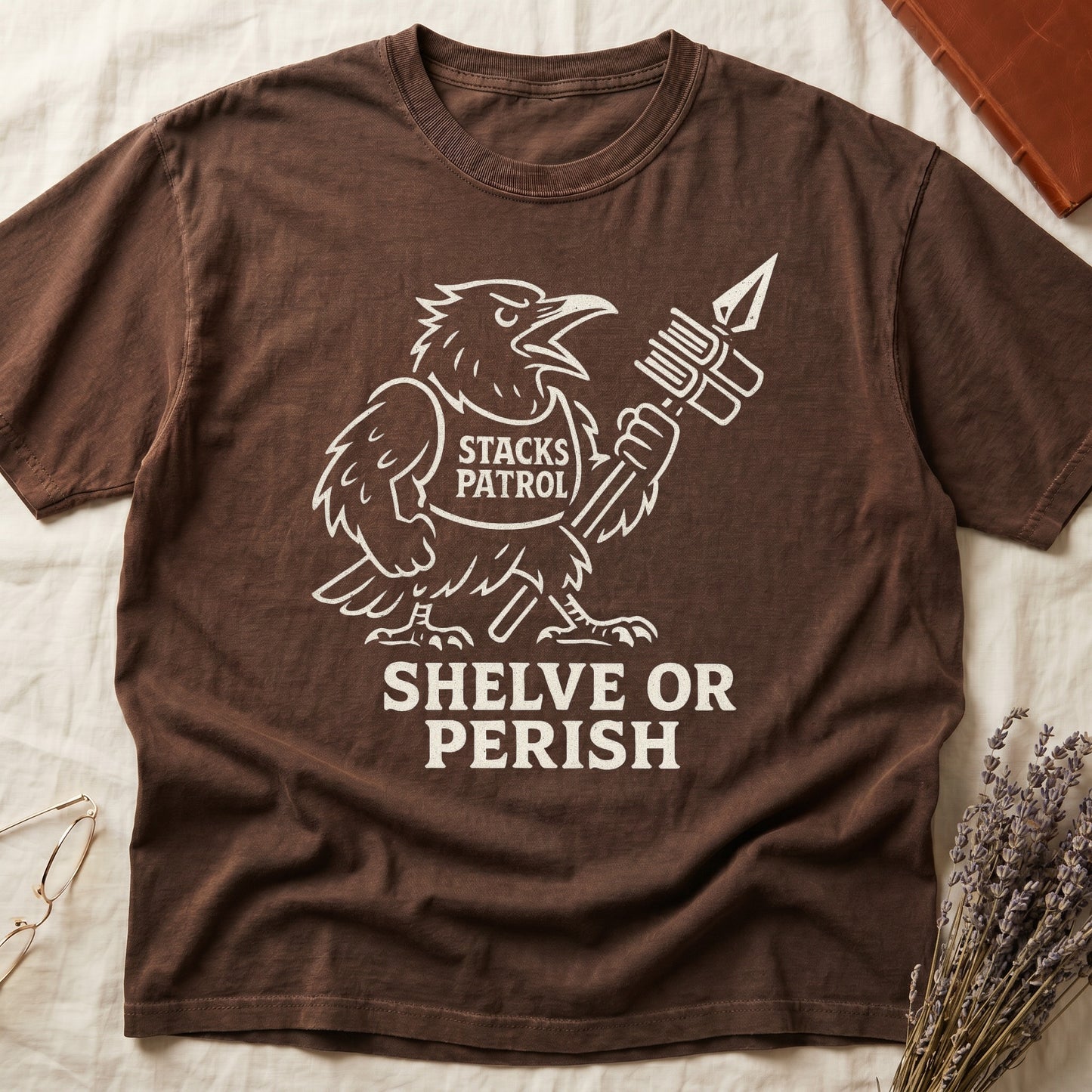 Espresso tee with vintage raven mascot, "STACKS PATROL" sash, feather pen aloft, "SHELVE OR PERISH" bold serif below.