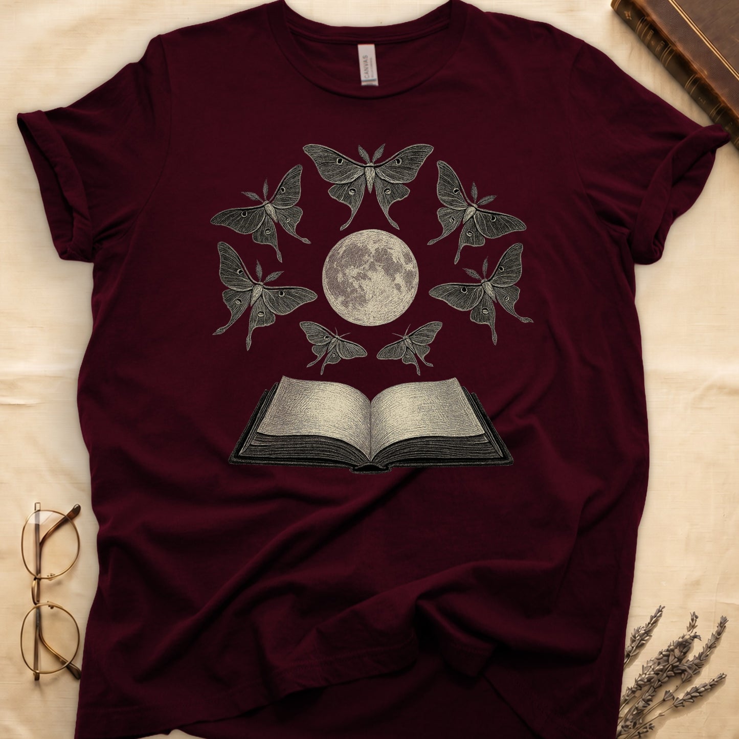 Maroon tee with full moon center, seven luna moths in halo around, open book below, cream-on-black engraving.