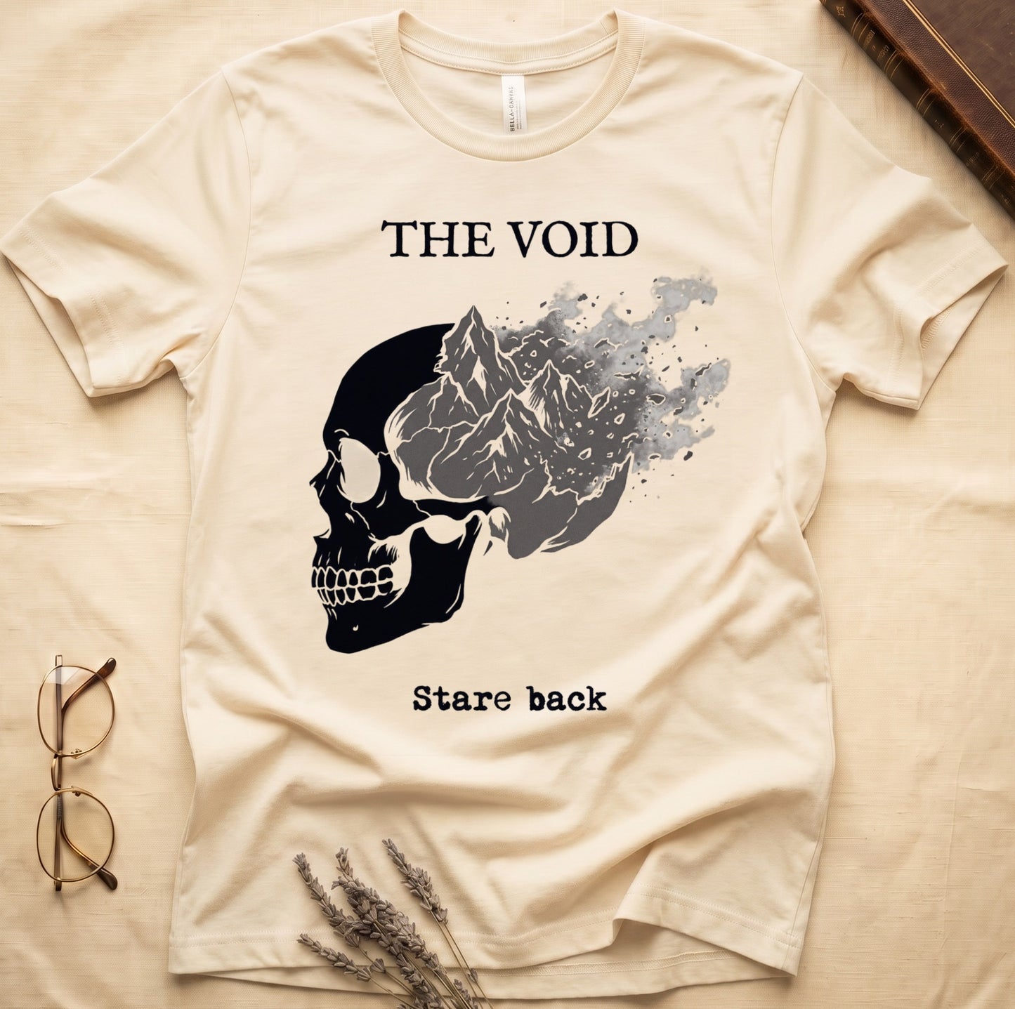 Natural tee with skull eroding into mountain landscape and stardust, "THE VOID" top, "Stare back" typewriter below.