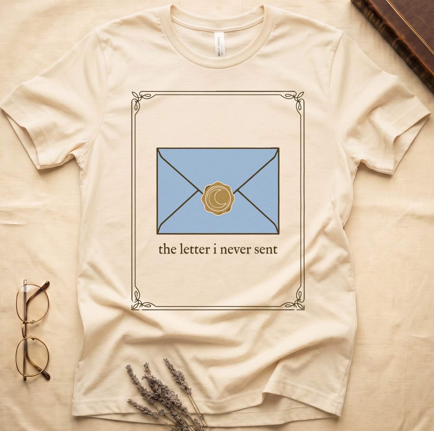 Natural tee with pale blue envelope sealed with gold crescent moon wax seal, "the letter i never sent" cream serif below.