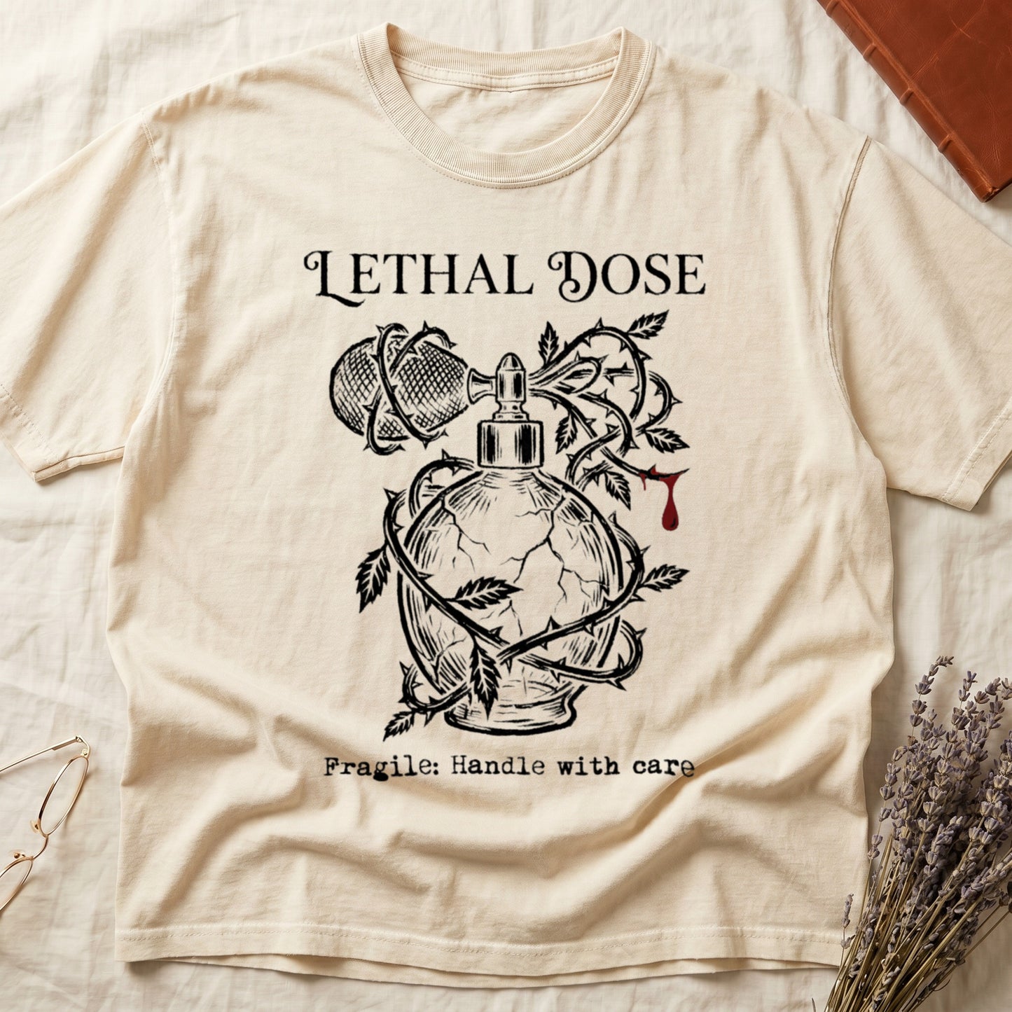 Ivory tee with cracked perfume bottle wrapped in thorns, blood drop, "LETHAL DOSE" top, "Fragile: Handle with care" below.