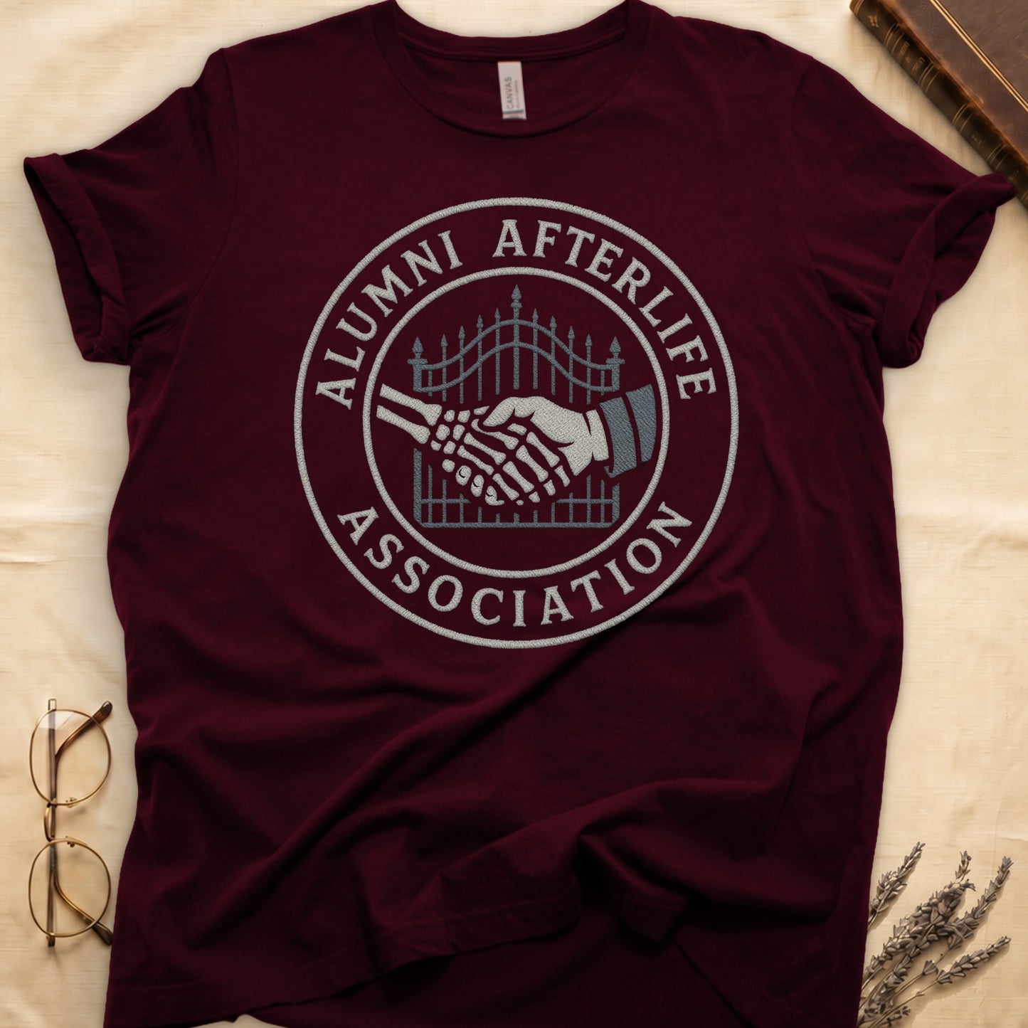 Navy tee with embroidered patch, skeletal handshake at cemetery gate, "ALUMNI AFTERLIFE" top, "ASSOCIATION" bottom.