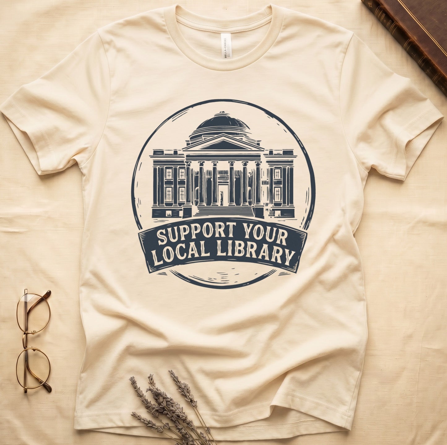 Natural tee with circular cream badge of a classical library building, "SUPPORT YOUR LOCAL LIBRARY" on a banner below.