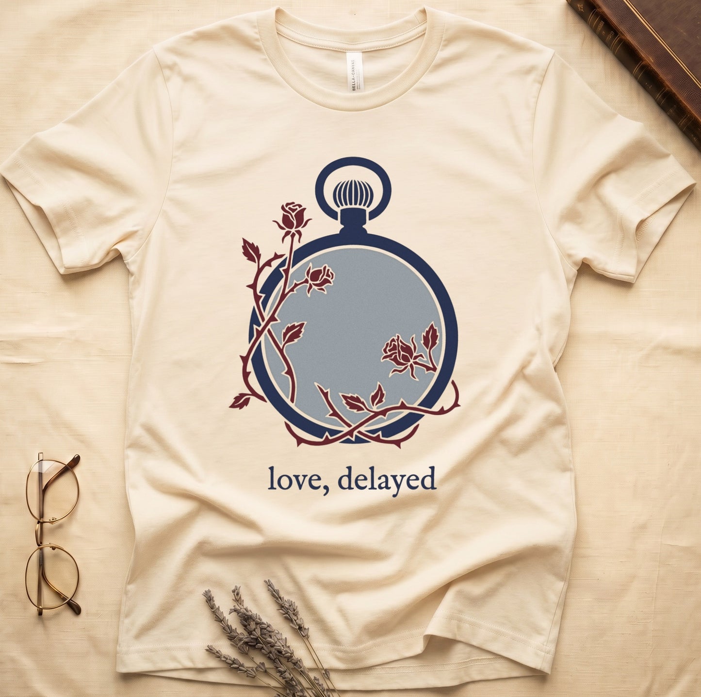 Natural tee with vintage cream pocket watch and dark red roses entwined, "love, delayed" in cream serif below.