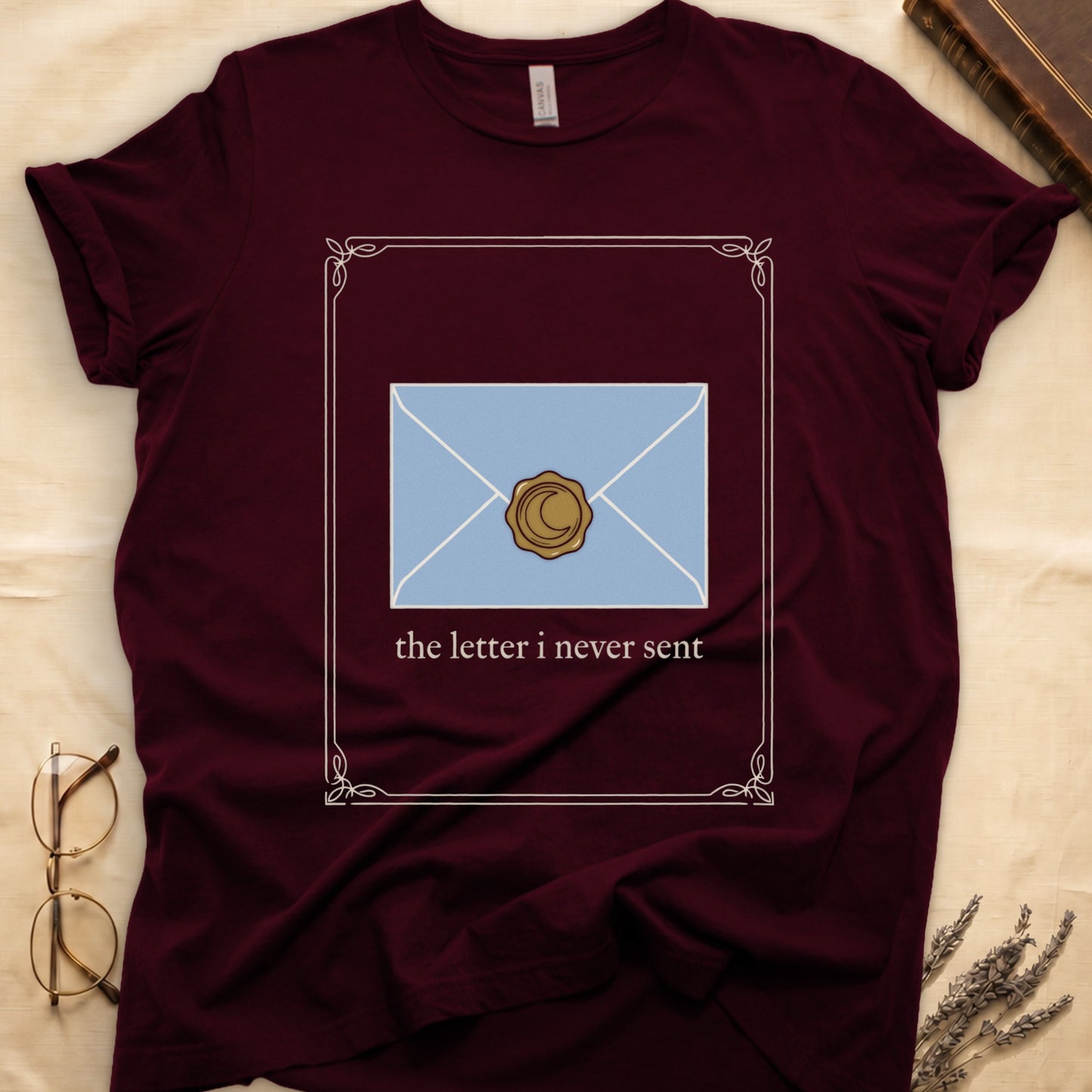 Maroon tee with pale blue envelope sealed with gold crescent moon wax seal, "the letter i never sent" cream serif below.