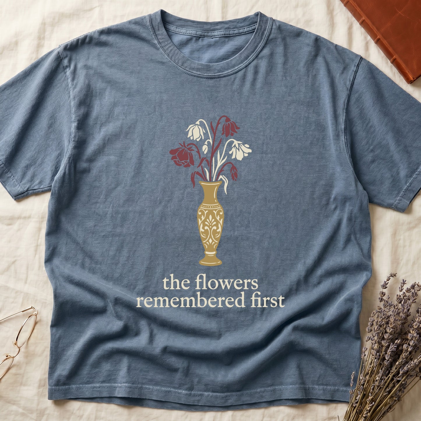 Blue Jean tee with gold ornate vase, wilting cream lilies and dark red roses, "the flowers remembered first" in cream serif.
