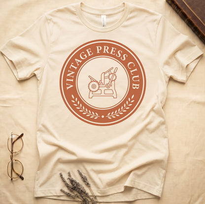 Natural tee with circular letterpress badge, "VINTAGE PRESS CLUB" in serif capitals, antique printing press in cream linework.