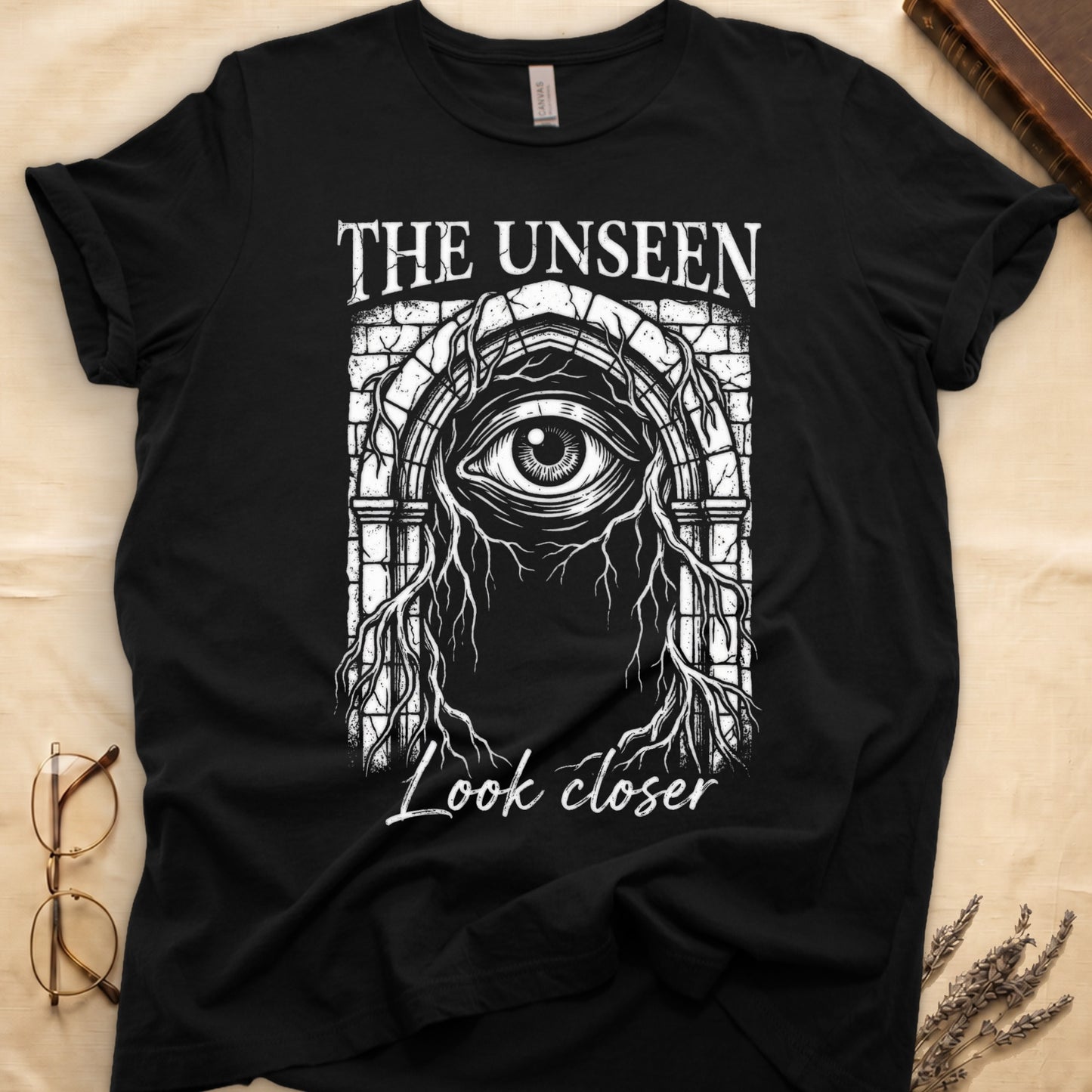 Black tee with Gothic stone arch, open eye, roots trailing, "THE UNSEEN" top, "Look closer" cursive below.