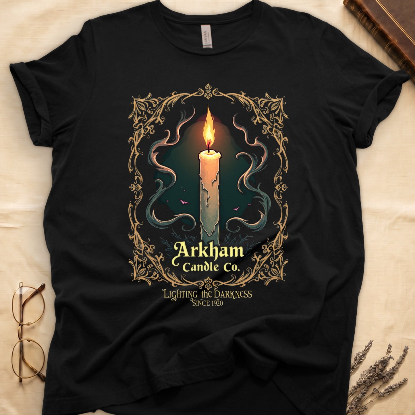 Black tee with lit candle, smoke, ornate gold filigree border, "Arkham Candle Co.," "Lighting the Darkness Since 1920."