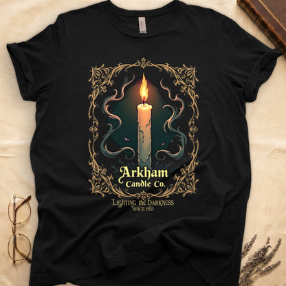 Black tee with lit candle, smoke, ornate gold filigree border, "Arkham Candle Co.," "Lighting the Darkness Since 1920."