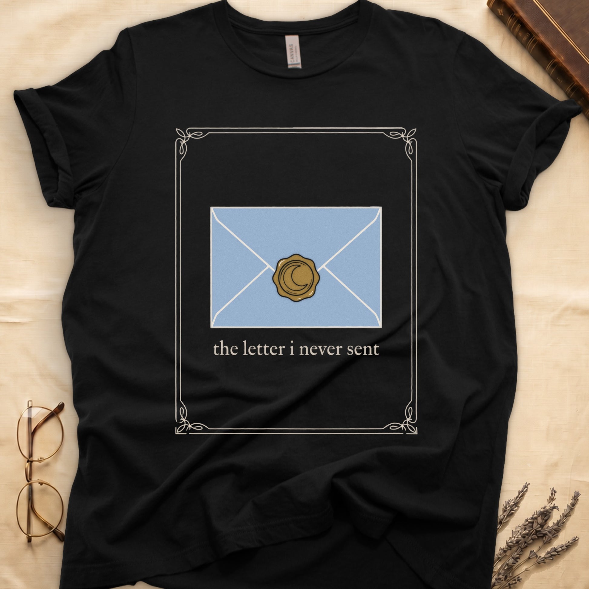 Black tee with pale blue envelope sealed with gold crescent moon wax seal, "the letter i never sent" cream serif below.