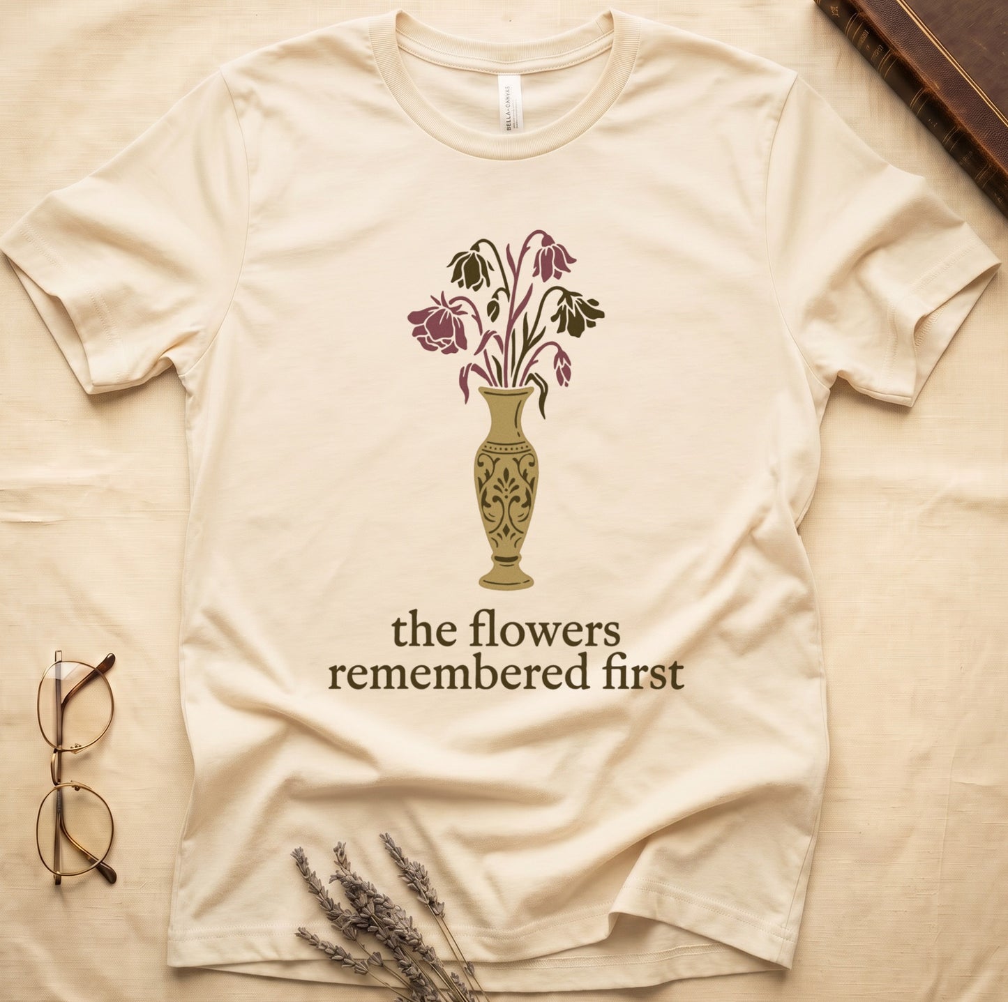 Natural tee with gold ornate vase, wilting cream lilies and dark red roses, "the flowers remembered first" in cream serif.