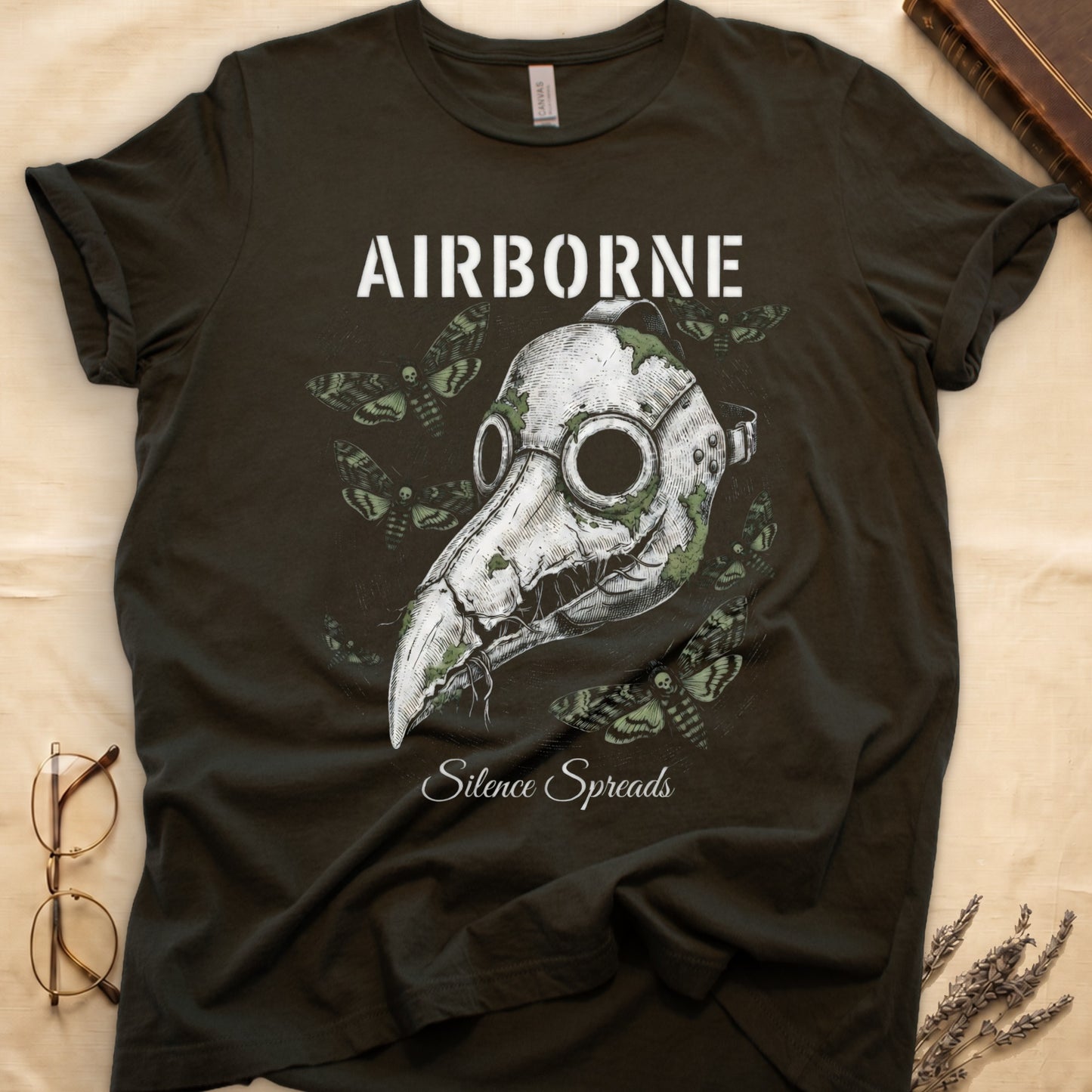 Military Green tee with plague doctor mask in cream linework, death's-head moths, "AIRBORNE" top, "Silence Spreads" cursive below.