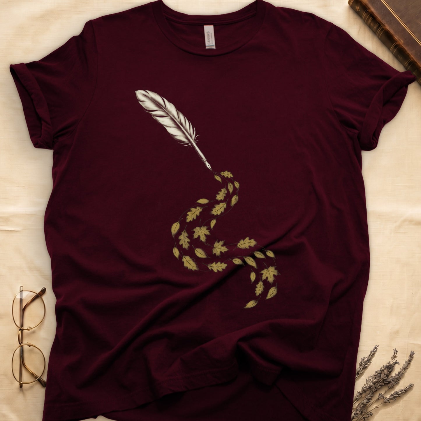 Maroon tee with antique quill pen in cream linework, oak and maple leaves cascading from the nib in gold and green.
