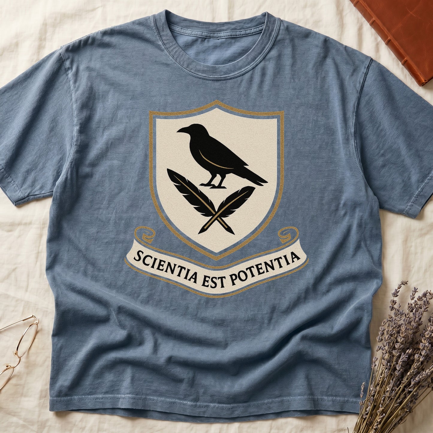 Blue Jean tee with cream-and-gold heraldic shield, black raven over crossed quills, scroll banner "SCIENTIA EST POTENTIA" below.