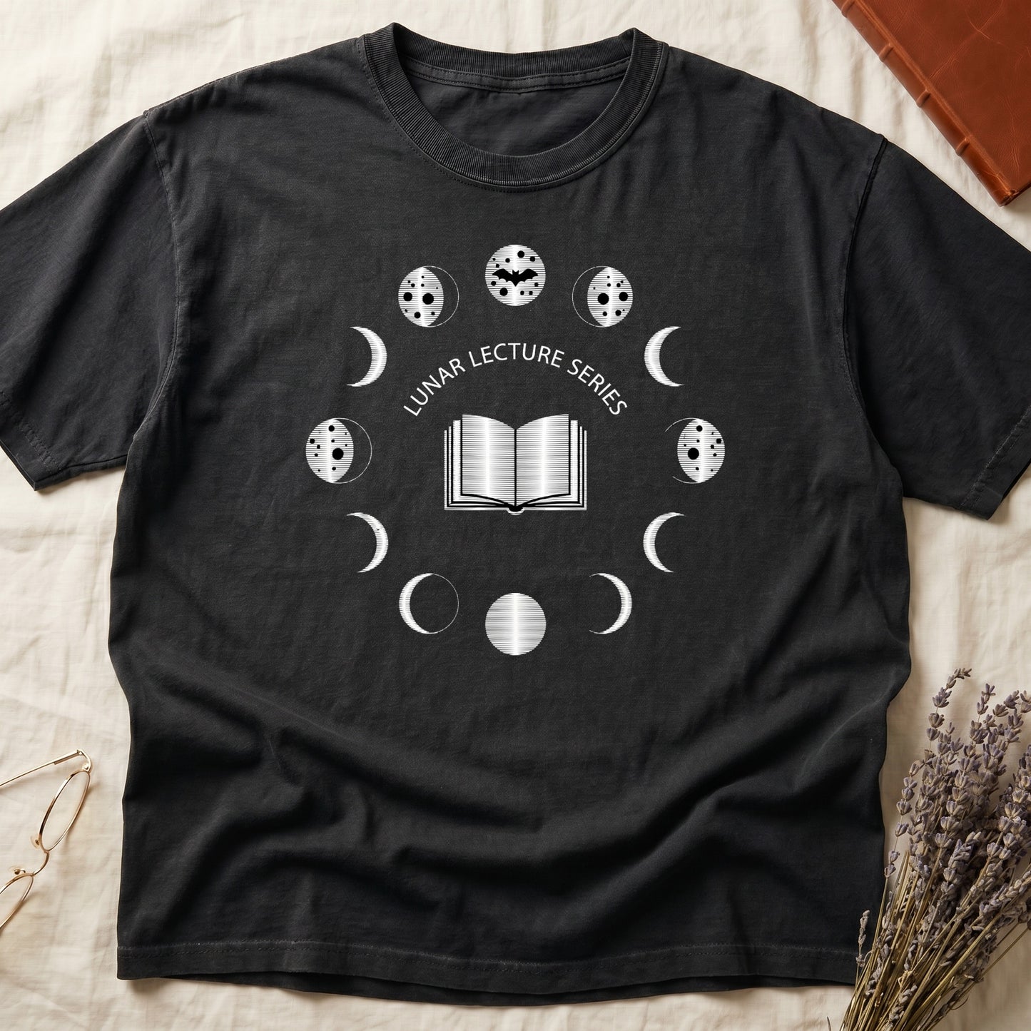 Black tee with open book at center, ring of cream moon phases around, curved "LUNAR LECTURE SERIES" double-arc above.