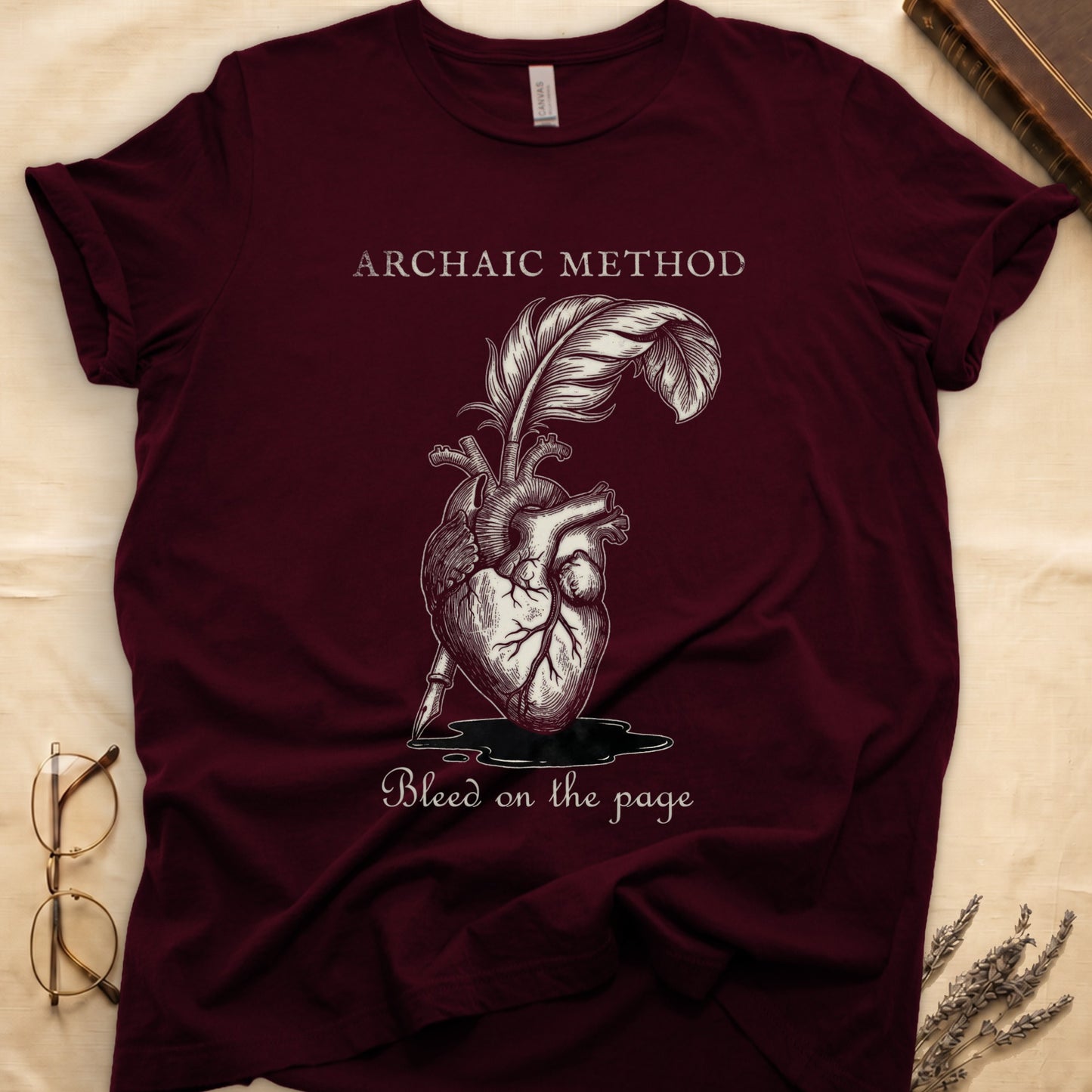 Maroon tee with anatomical heart and quill emerging in cream linework, "ARCHAIC METHOD" top, "Bleed on the page" below.