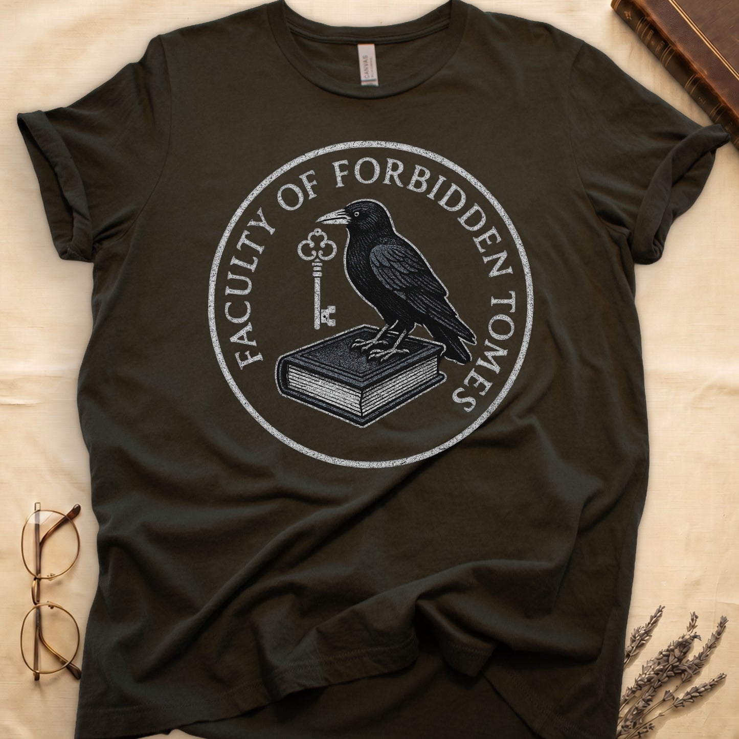 Maroon tee with circular badge, raven on closed book holding key, "FACULTY OF FORBIDDEN TOMES" curved around in cream.