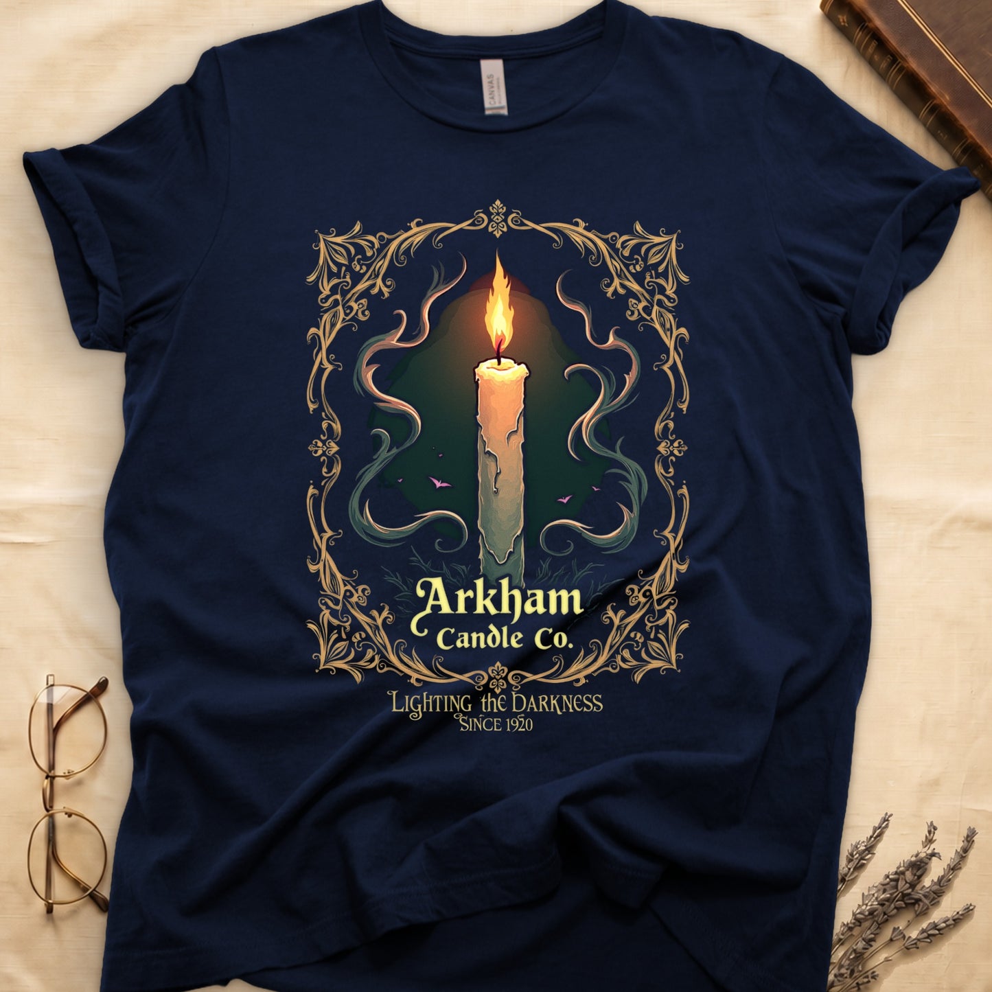 Navy tee with lit candle, smoke, ornate gold filigree border, "Arkham Candle Co.," "Lighting the Darkness Since 1920."