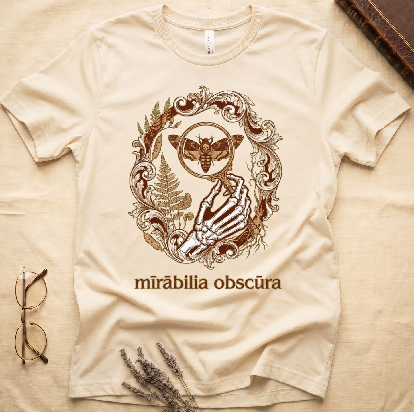 Natural tee with skeletal hand, magnifying glass, death's-head moth, ferns on green, "mīrābilia obscūra" in Latin italic.