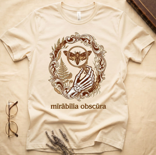 Natural tee with skeletal hand, magnifying glass, death's-head moth, ferns on green, "mīrābilia obscūra" in Latin italic.