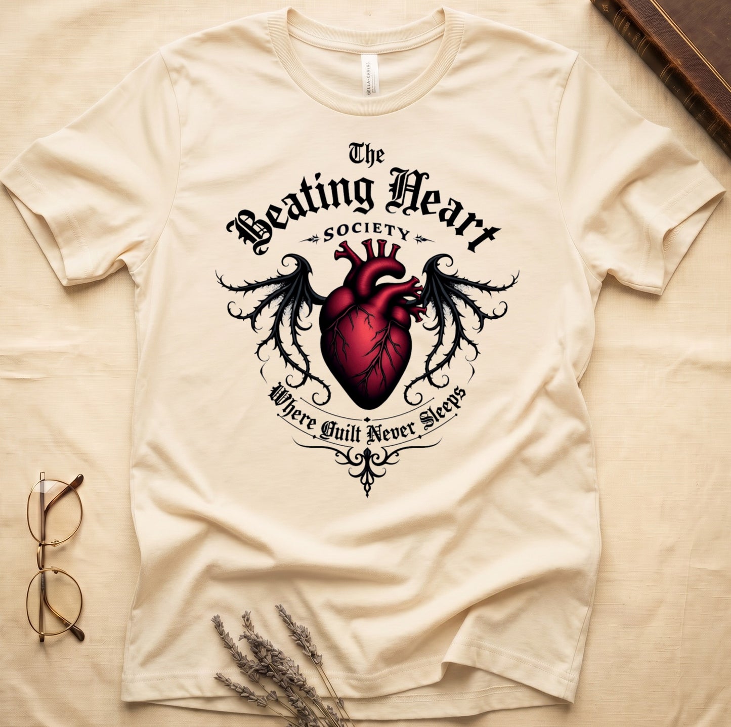 Natural tee with anatomical red heart, gothic wings, "The Beating Heart SOCIETY," "Where Guilt Never Sleeps" below.