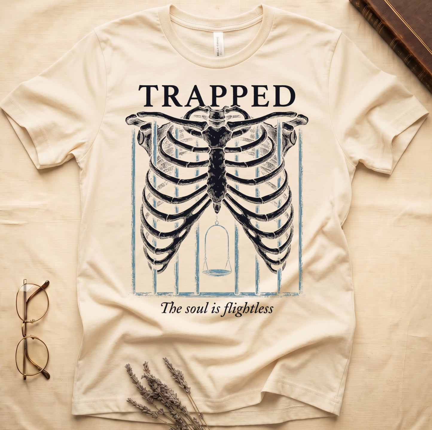 Natural tee with anatomical ribcage on prison bars, bird swing inside, "TRAPPED" top, "The soul is flightless" italic below.