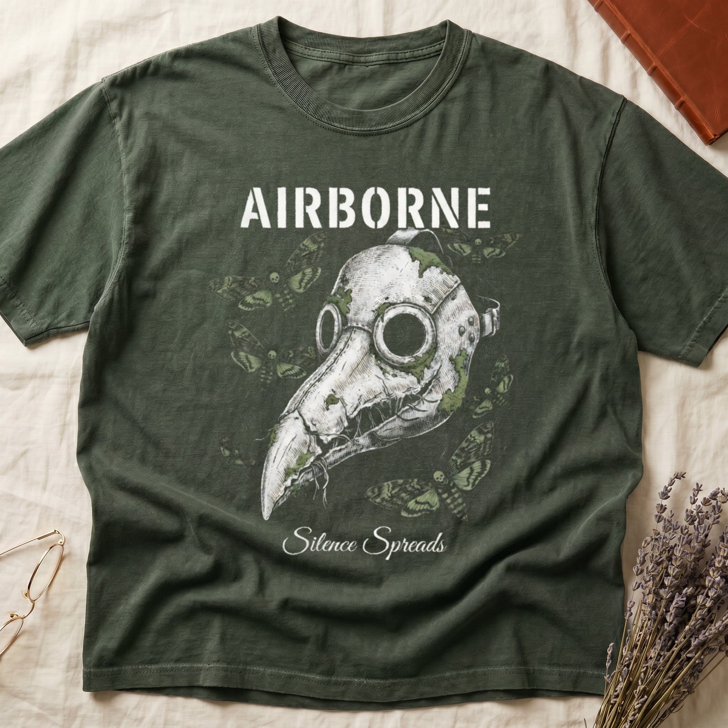 Green tee with plague doctor mask in cream linework, death's-head moths, "AIRBORNE" top, "Silence Spreads" cursive below.