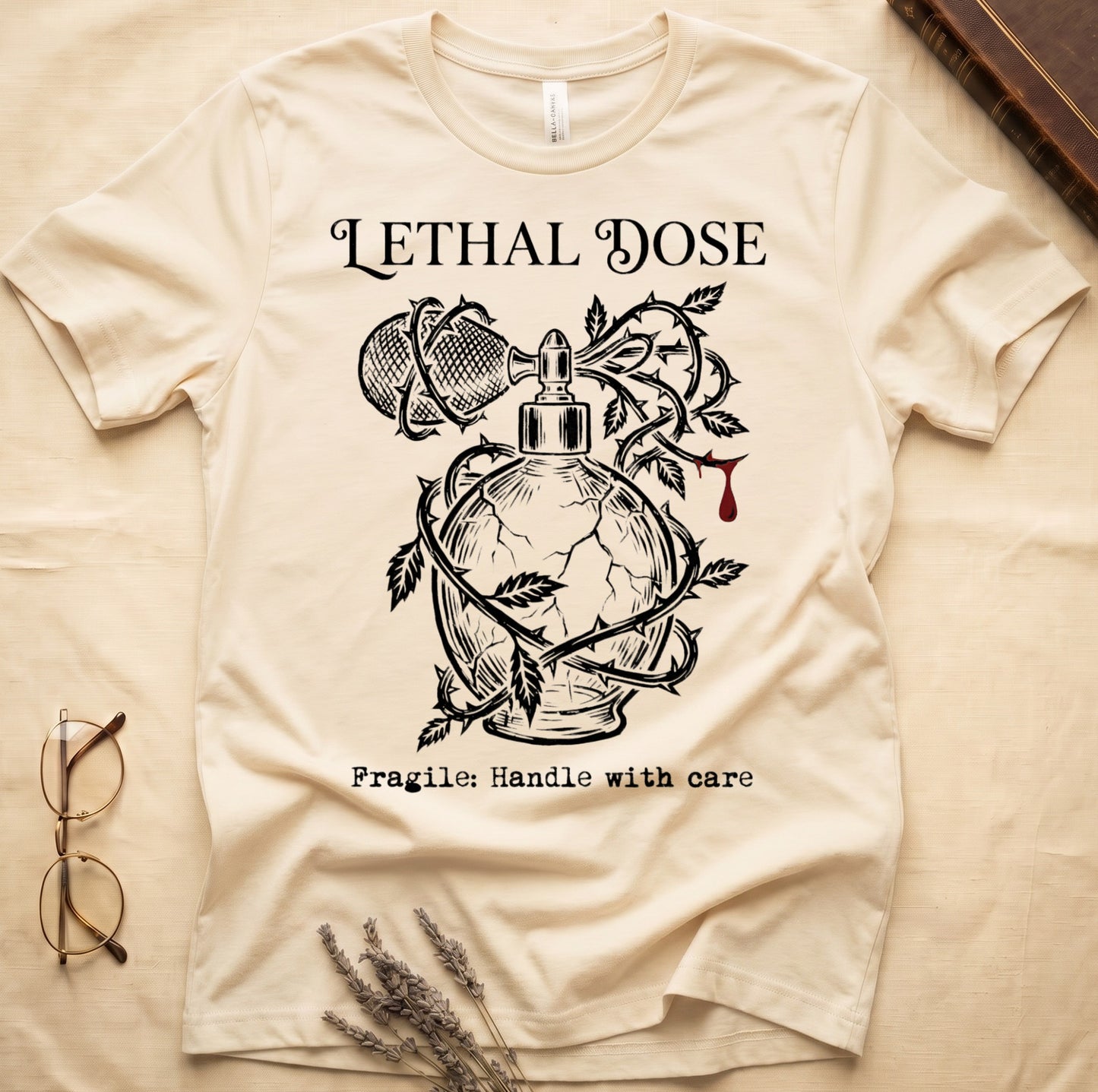 Natural tee with cracked perfume bottle wrapped in thorns, blood drop, "LETHAL DOSE" top, "Fragile: Handle with care" below.