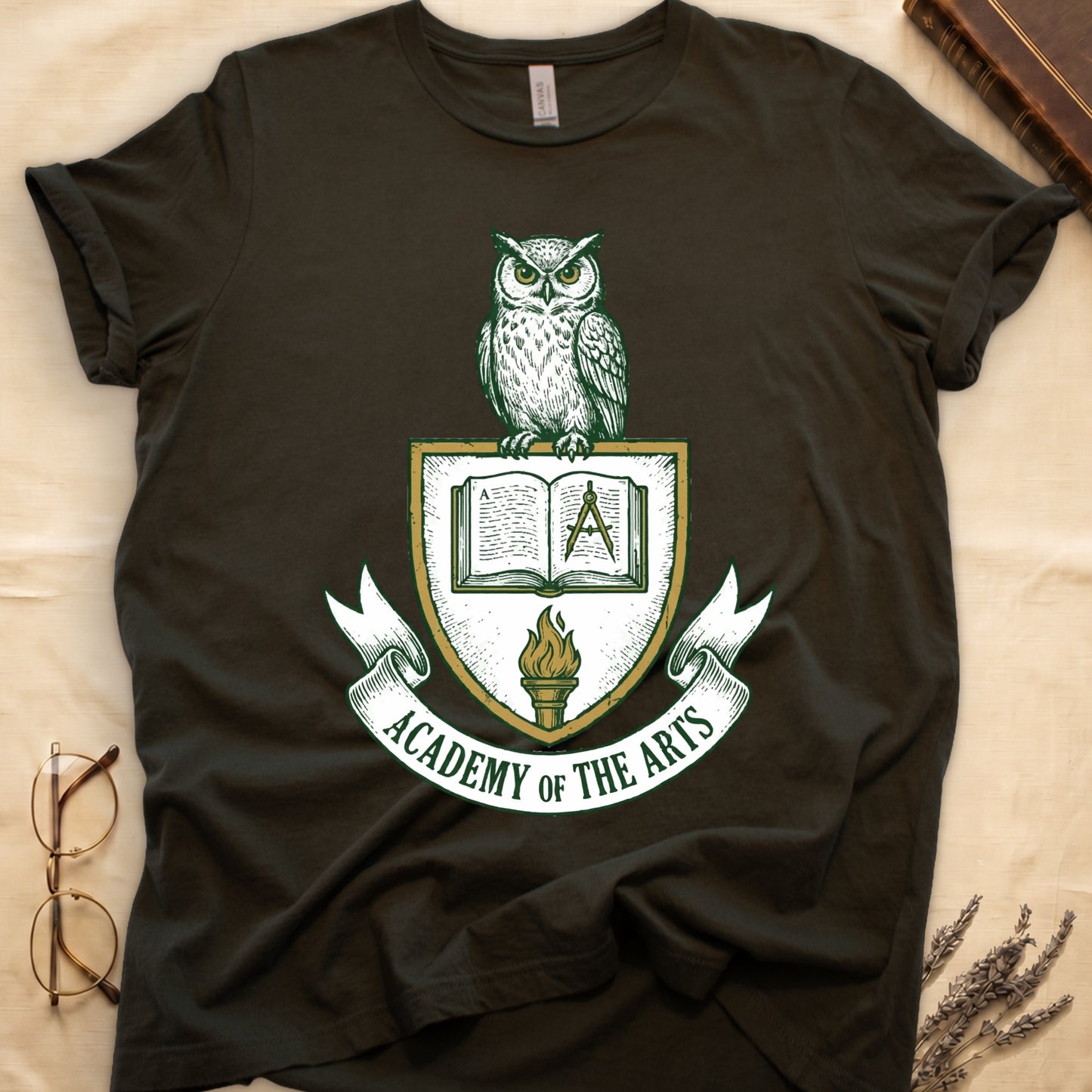 Military Green tee with heraldic shield crest, owl perched on top, open book and dividers center, "ACADEMY OF THE ARTS" on a banner.