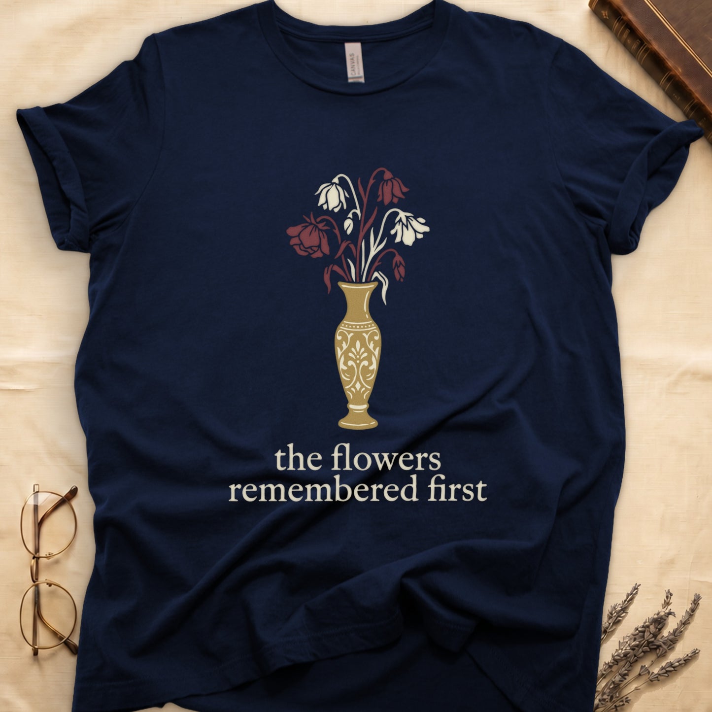 Navy tee with gold ornate vase, wilting cream lilies and dark red roses, "the flowers remembered first" in cream serif.