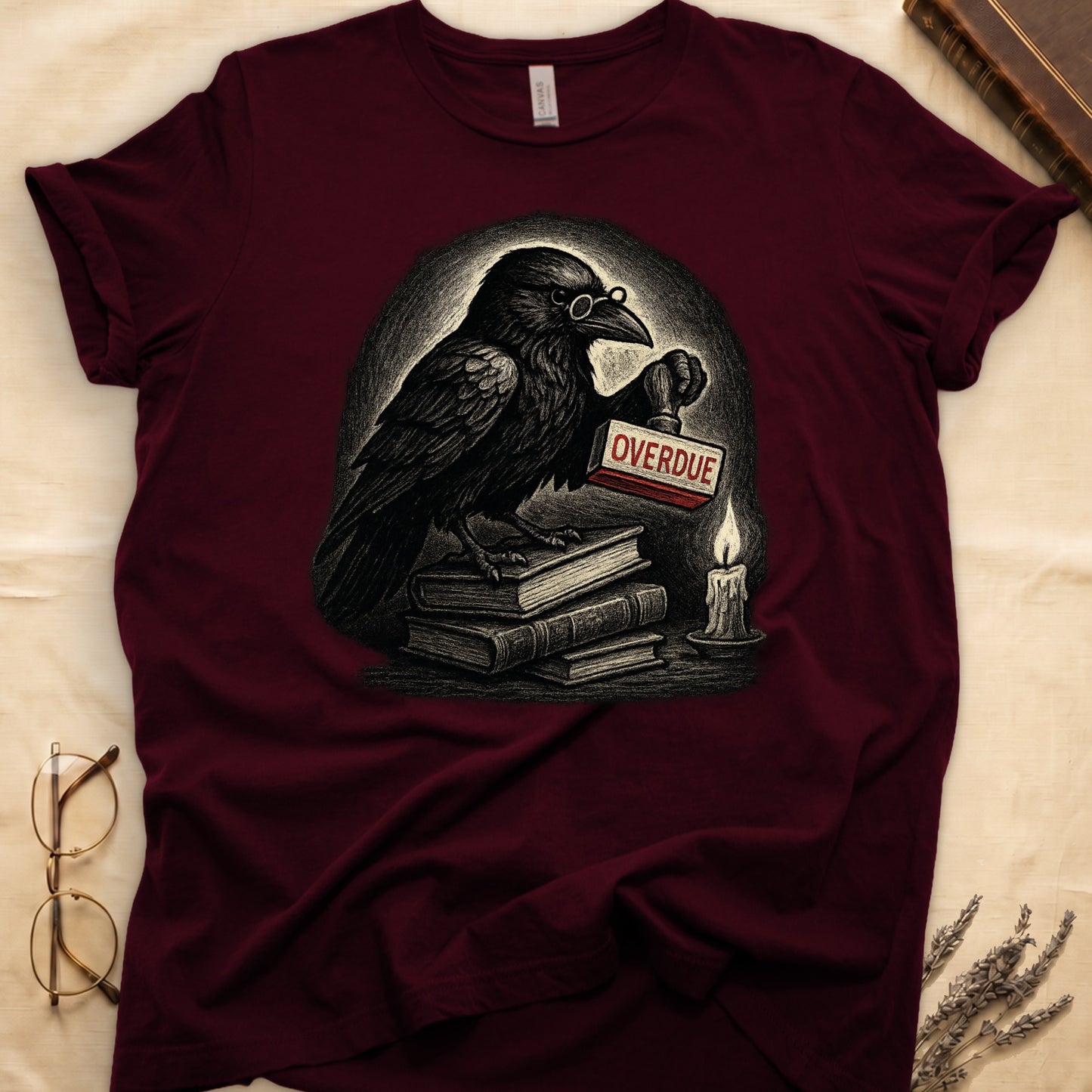 Maroon tee with bespectacled raven on stacked books, holding wooden "OVERDUE" stamp, lit candle beside, dawn glow behind.