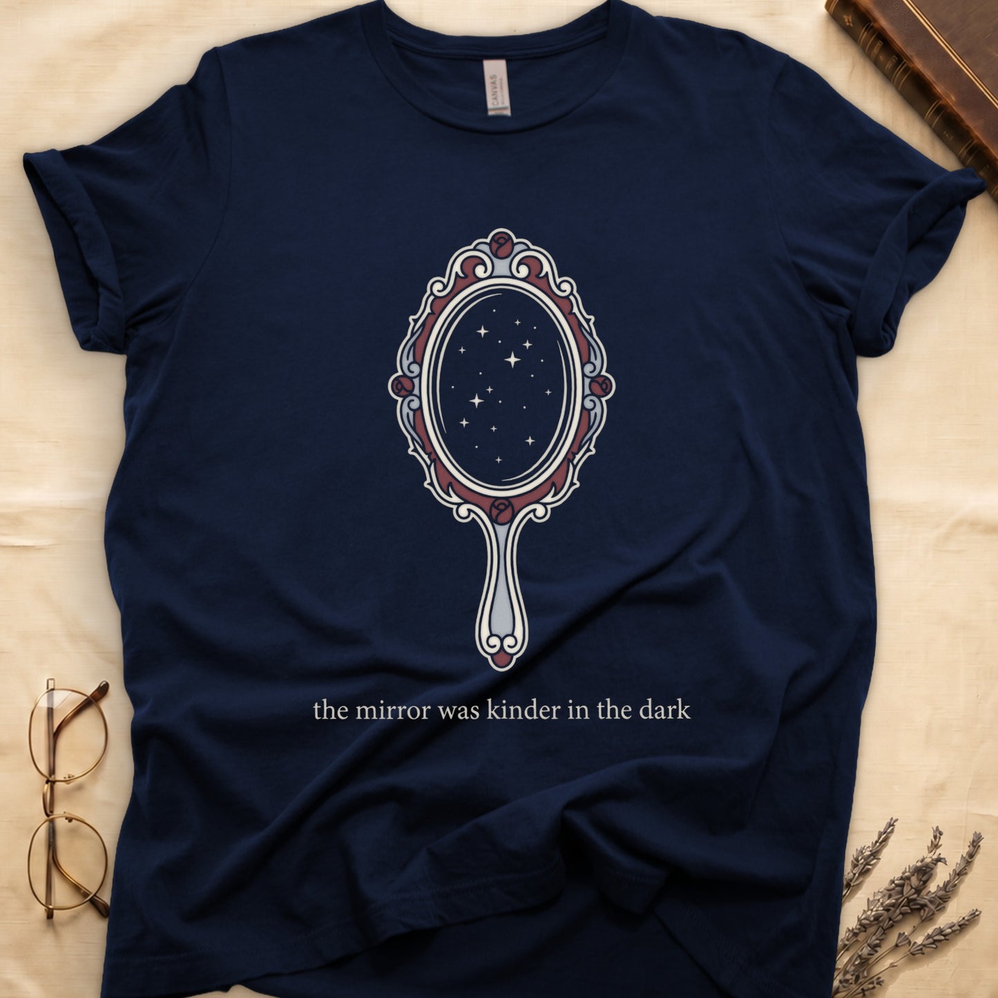 Navy tee with ornate hand mirror, dark red roses, starfield reflection, "the mirror was kinder in the dark" serif below.