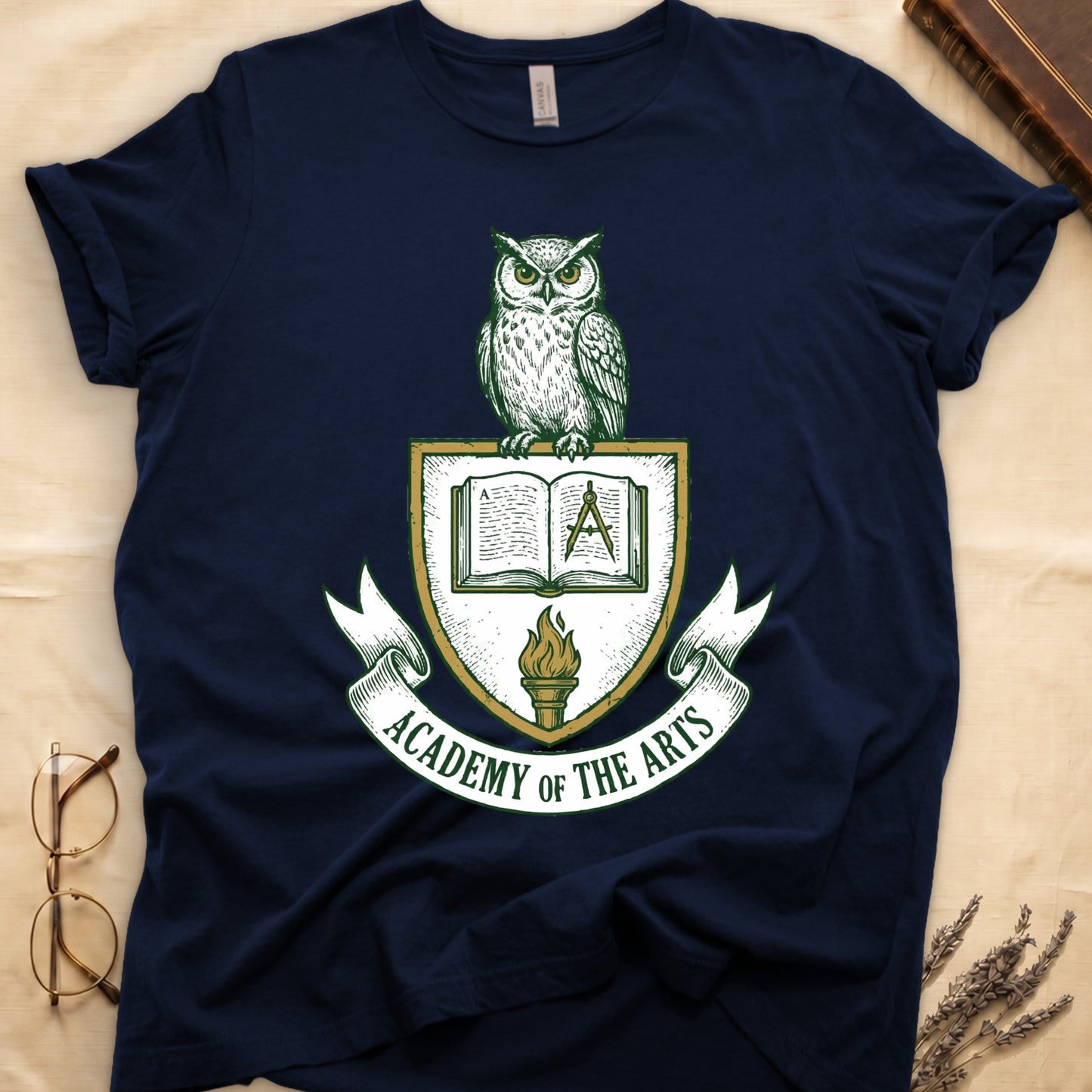 Navy tee with heraldic shield crest, owl perched on top, open book and dividers center, "ACADEMY OF THE ARTS" on a banner.