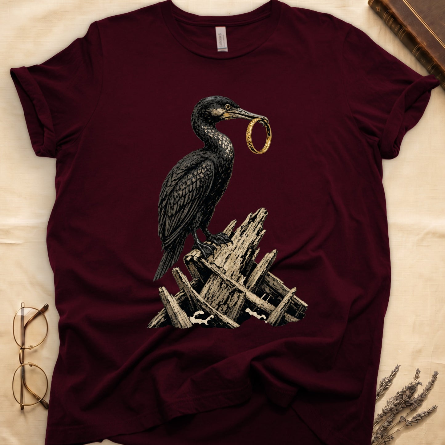 Maroon t-shirt with a crow holding a gold ring perched on weathered wood.