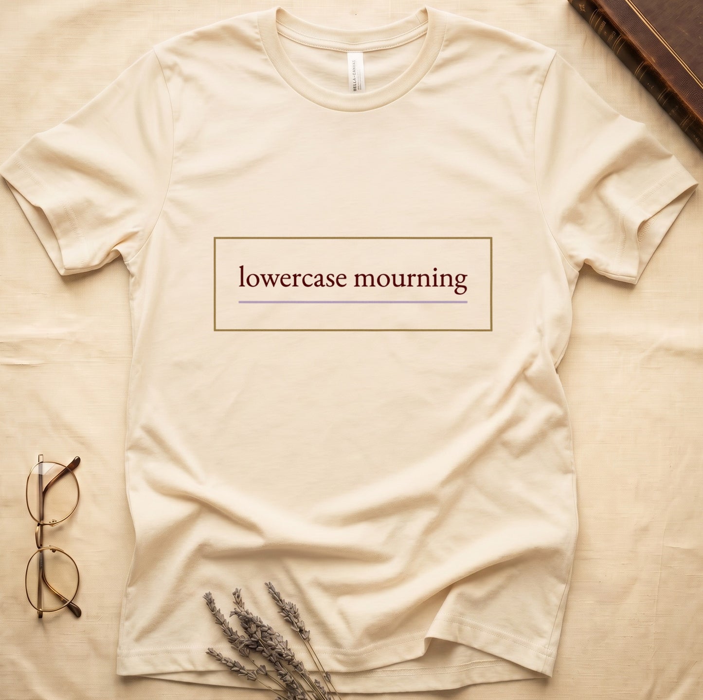Natural tee with "lowercase mourning" in cream serif inside gold rectangle frame, lavender line below.