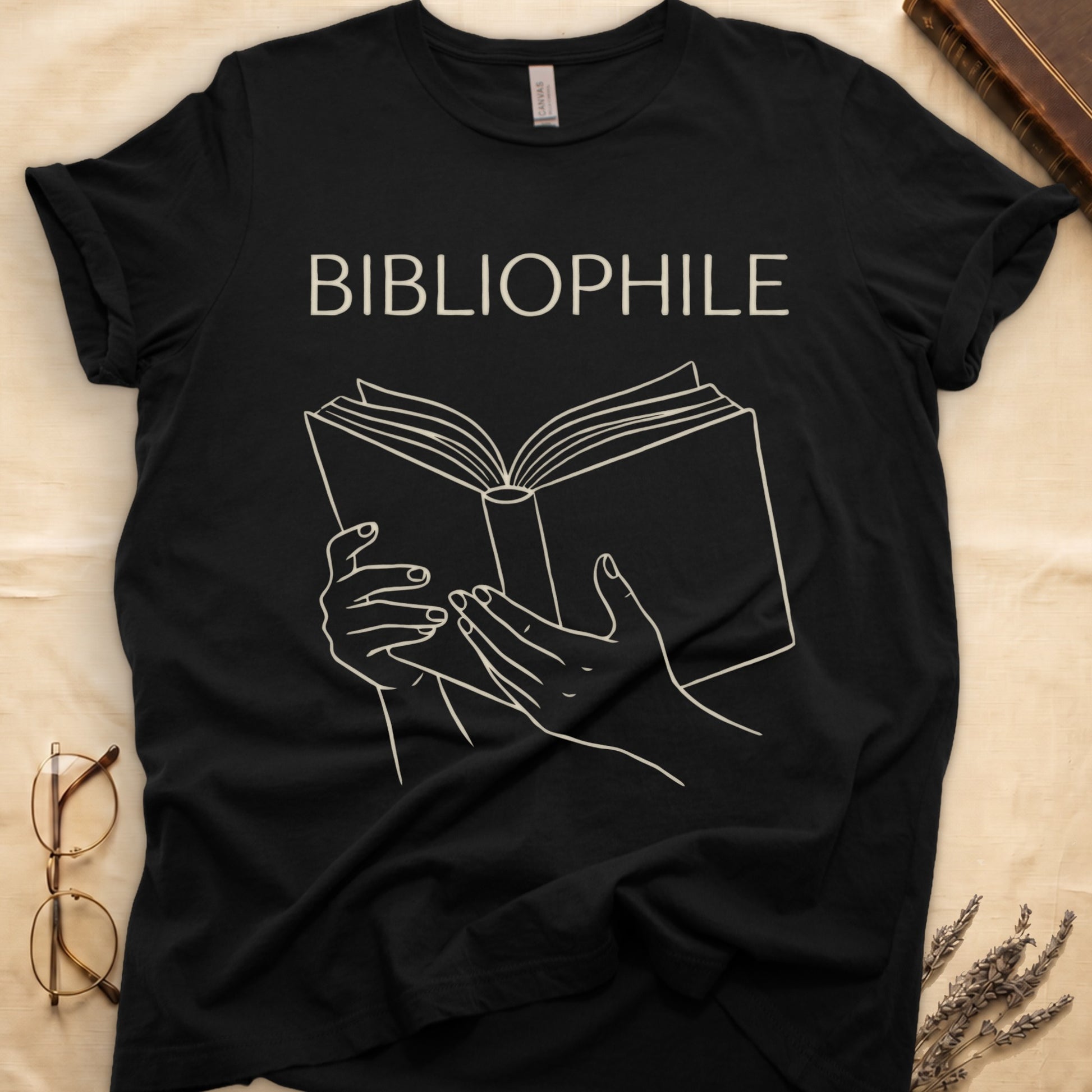 Black tee with two hands cradling an open book in cream and brown linework, "BIBLIOPHILE" in serif capitals above.