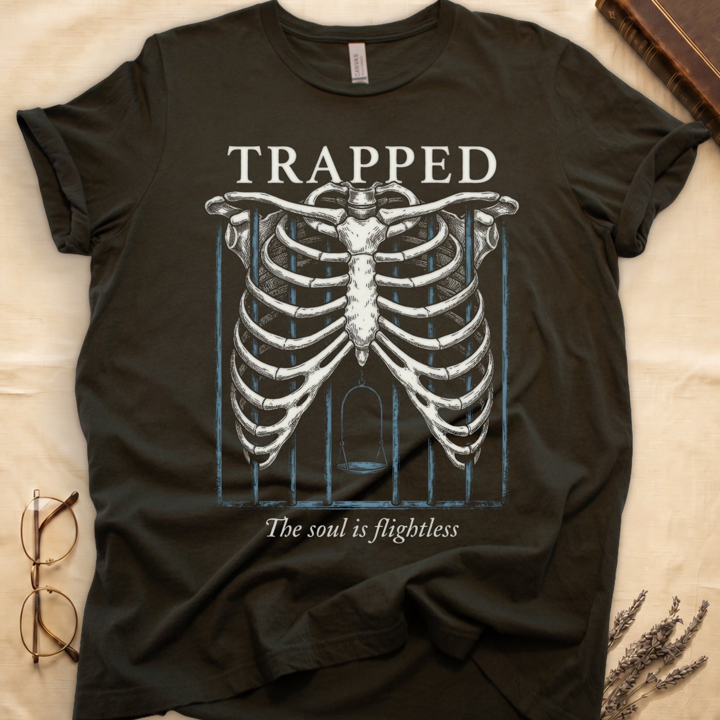 Military Green tee with anatomical ribcage on prison bars, bird swing inside, "TRAPPED" top, "The soul is flightless" italic below.