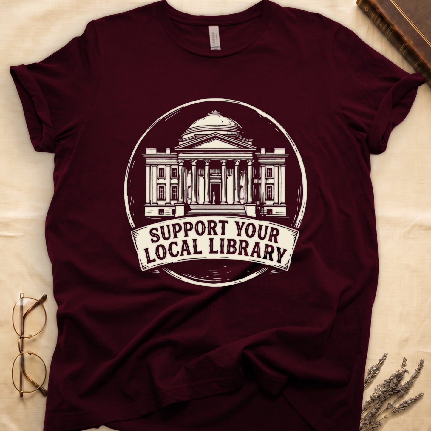 Maroon tee with circular cream badge of a classical library building, "SUPPORT YOUR LOCAL LIBRARY" on a banner below.