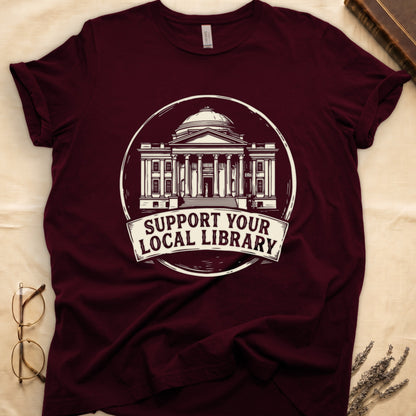 Maroon tee with circular cream badge of a classical library building, "SUPPORT YOUR LOCAL LIBRARY" on a banner below.