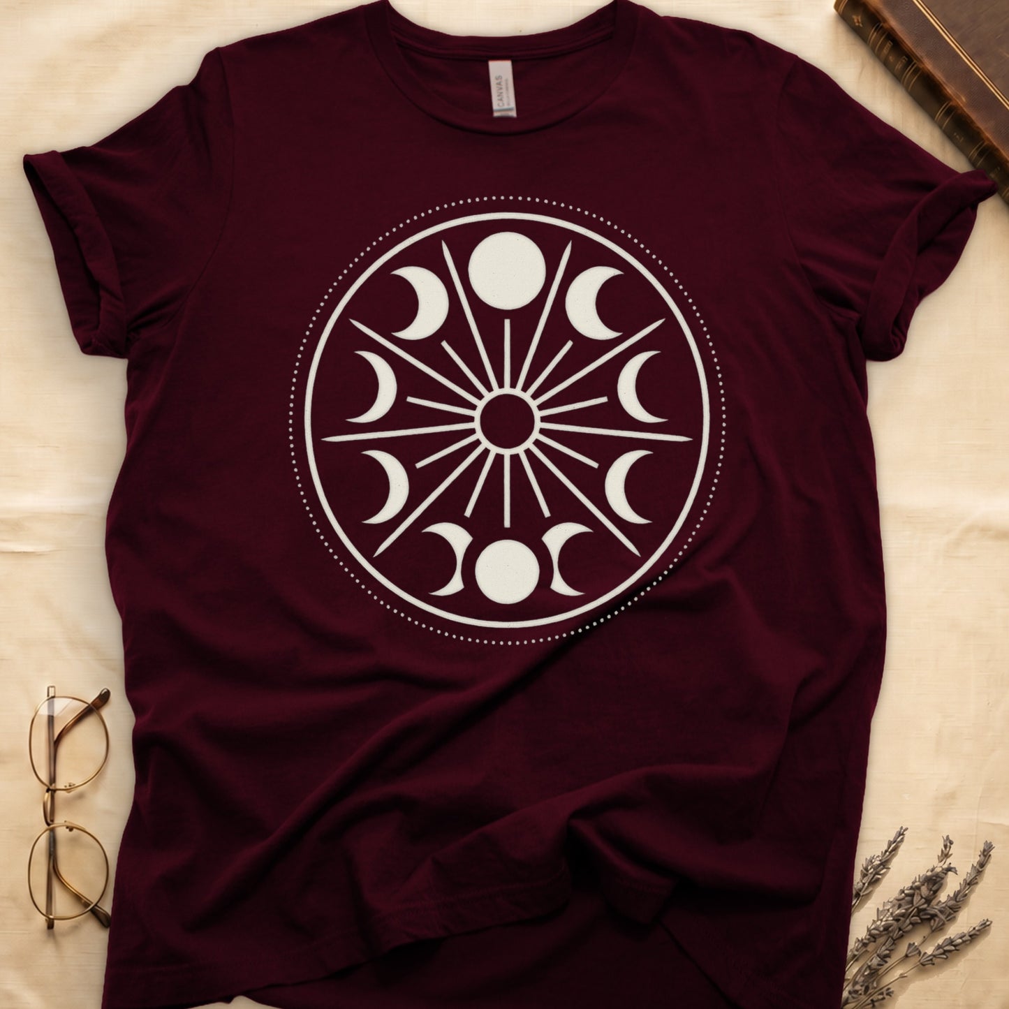 Maroon tee with cream lunar orrery wheel, dotted ring, radial sunburst, moon phases around perimeter, central star.