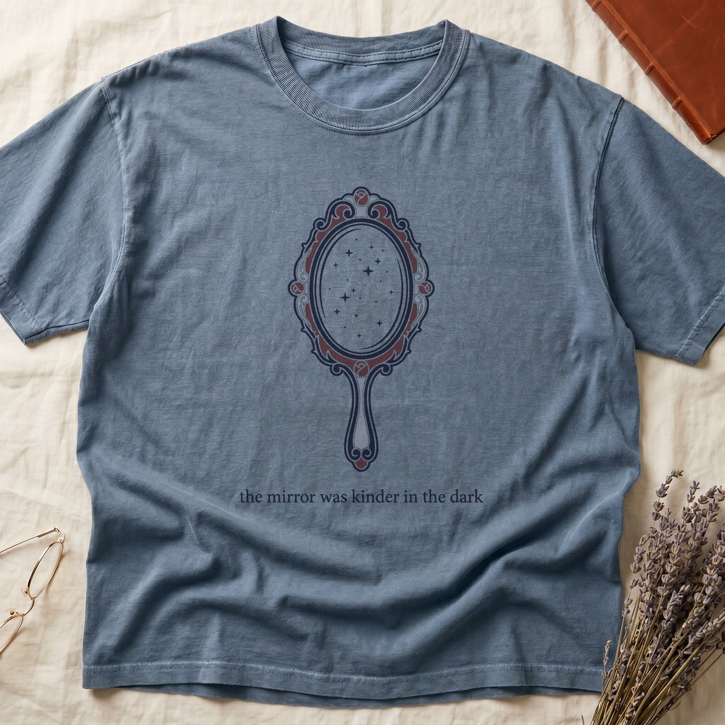 Blue Jean tee with ornate hand mirror, dark red roses, starfield reflection, "the mirror was kinder in the dark" serif below.