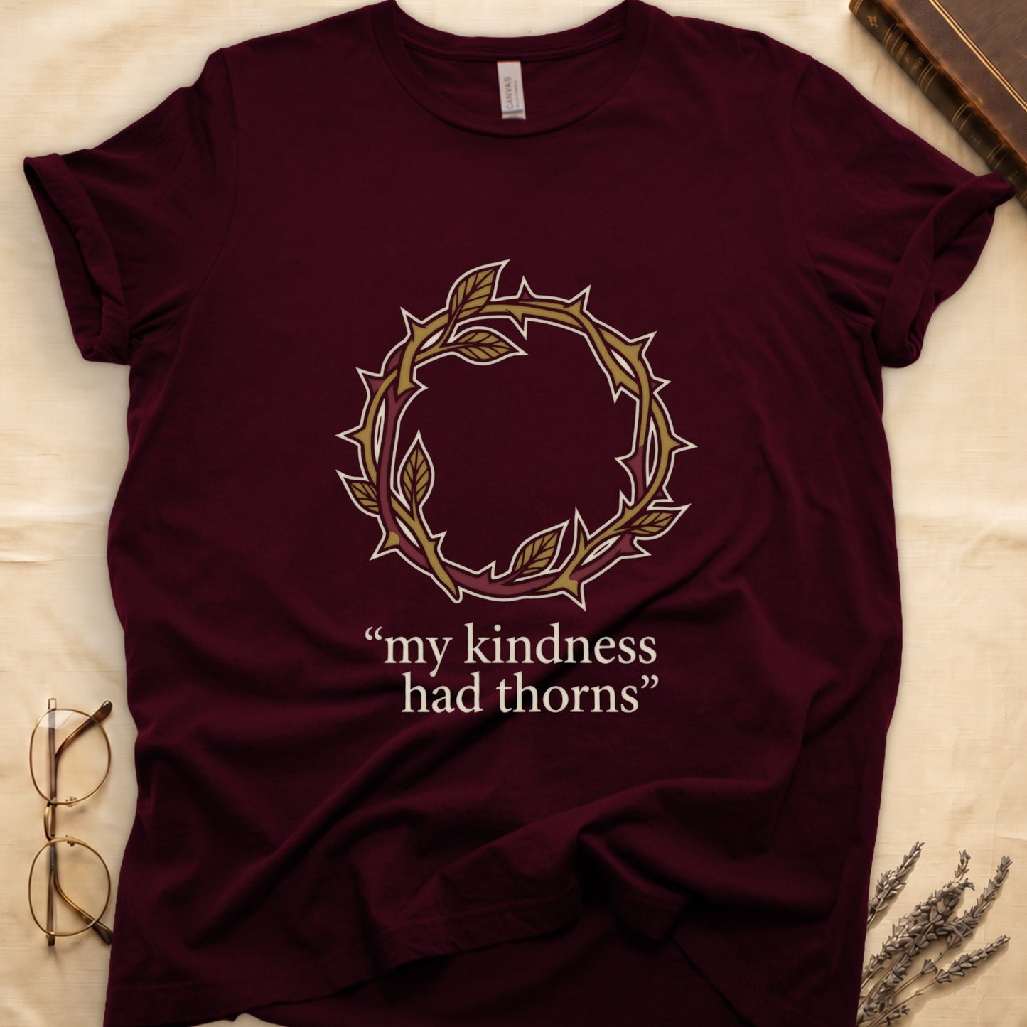 Maroon tee with crown of thorns in gold and red with green leaves, "my kindness had thorns" in cream serif.